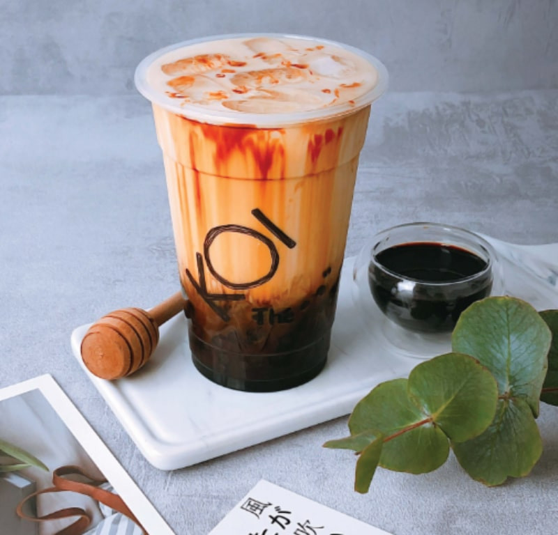 Một ly trà sữa Koi Thé cỡ lớn với trân châu vàng, được đặt trên một mặt phẳng màu trắng