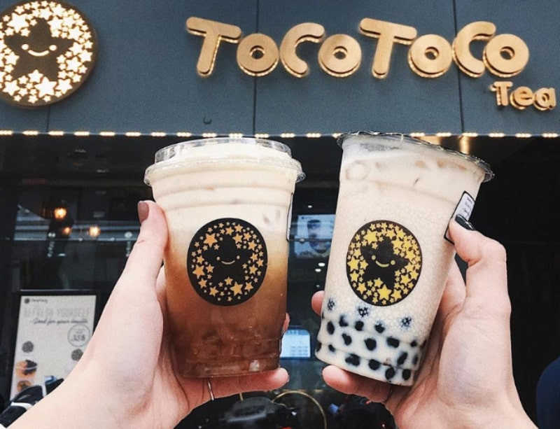 Một ly trà sữa TocoToco với trân châu và thạch, đặt trên bàn gỗ cùng logo thương hiệu