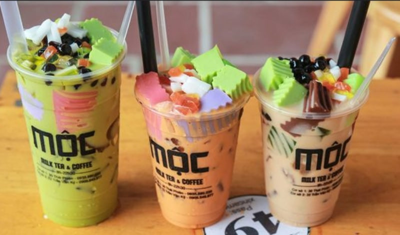 Một ly trà sữa Trà Sữa Mộc với lớp kem sữa và bột trà xanh rắc lên, đặt trên bàn đá